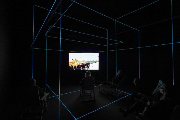 Hito Steyerl - Factory of the Sun - Eenkanaals HD-video met omgevingssculptuur, lichtgevend LED-raster, strandstoelen kleur en geluid, 23 minuten, 2015